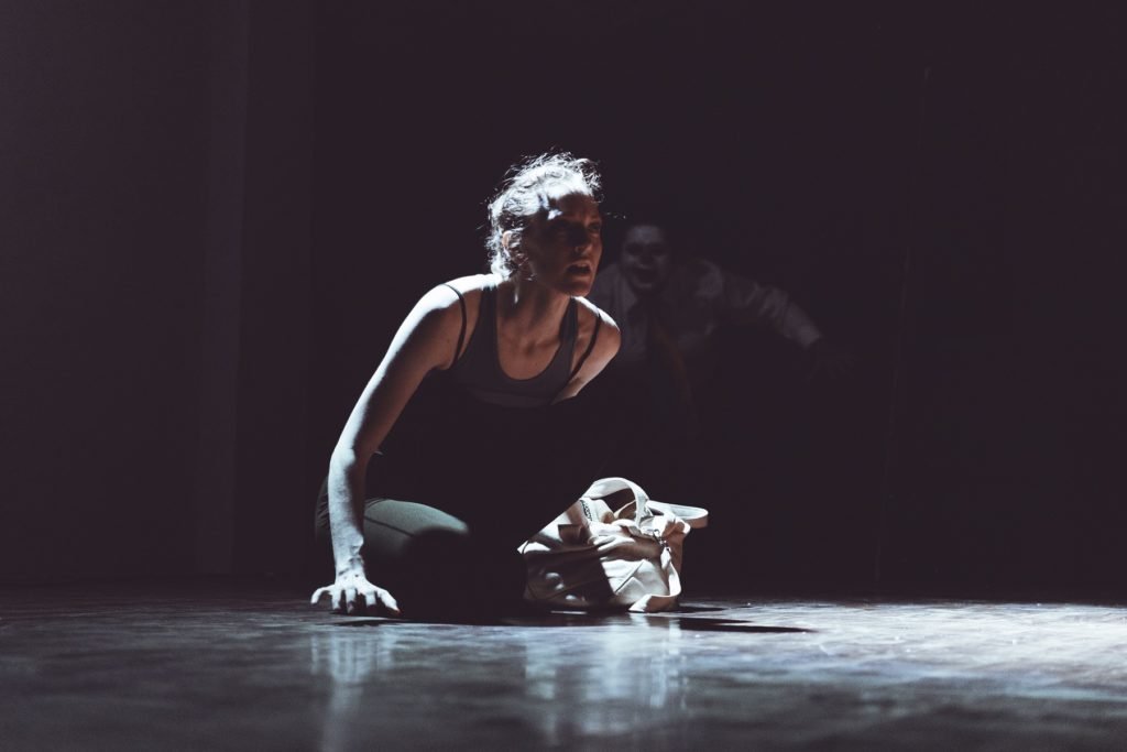 Gillian sitting in a spotlight on a dark stage looking frightened